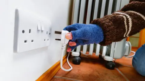 Getty Images A woman wearing a blue fingerless glove and a brown woollen jumper plugs a portable electric heater into a mains socket.