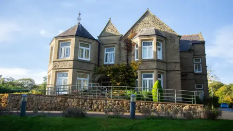 Parkland Place Rehabilitation Centre in north Wales is a large 16-bed unit surrounded by gardens. The sky is blue with the sun shining across the brickwork of the large two-storey building. A sloped entrance is at the front of the property where guests enter.