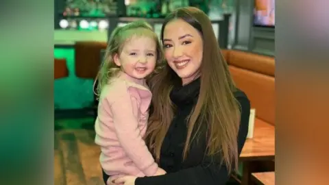 Laura Maguire Laura poses while holding a small child with blonde hair. They are standing in front of a table at a restaurant and are smiling at the camera. Laura is a young woman with long, brown hair which has been straightened. She is wearing a black top. The girl is wearing a light pink knitted dress.