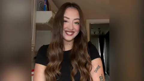 Laura Maguire Laura, a young woman, takes a selfie in a kitchen. She has long, brown hair and is smiling at the camera. She is wearing a black t-shirt and has two, small tattoos on her arm.