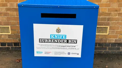 A large blue bin with a slot and a sign saying 'Knife Surrender Bin on it'. The bin is positioned in front of a brick wall.