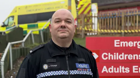 A police officer in his black uniform standing next to a hospital sign, with an ambulance in the background behind him.