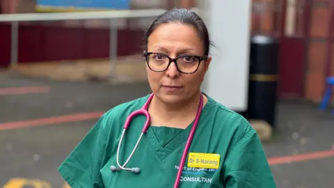 A woman in green medical scrubs with a stethoscope around her neck looks at the camera with a solemn expression. Her yellow name badge says 'Dr S Narang'.