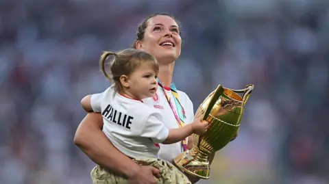 Getty Images Abbie Ward, wearing her white England shirt, is pictured carrying her daughter Hallie, a toddler who has her hair in pigtails. Both are carrying the gold trophy.