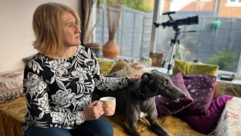 Kate Roux, with short blonde hair and wearing a black top with a white floral print, is sitting on a sofa holding a cup of tea and stroking a black whippet dog as she looks out of her conservatory window