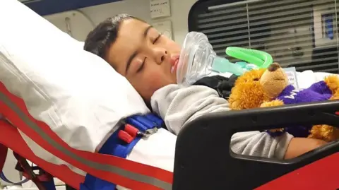 Family handout A boy with dark hair lies on a stretcher in an ambulance with a tube in his mouth. He is unconscious. A teddy bear lies next to him. 