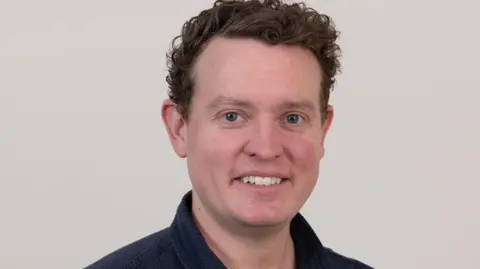UKHSA A middle aged man in a dark blue shirt is smiling into the camera in front of a neutral grey background. 