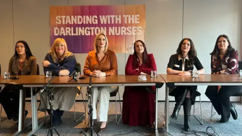 Six women sit at a long table with microphones set up in front of them. Behind them is a banner with the words 