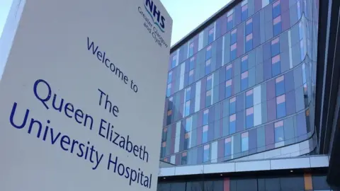 A picture of the Queen Elizabeth University Hospital taken from below, looking up to the grey, purple and pink building. An NHS sign with the hospital's name sits in the foreground.
