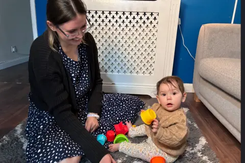 Chloe Chloe sits on a grey and cream rug with her daughter who is playing with colourful stacking cups and looking over at the camera, wearing a knitted beige jumper and leggings with a floral pattern. Chloe is wearing a black and white dress with a black cardigan, smiling down at her daughter.
