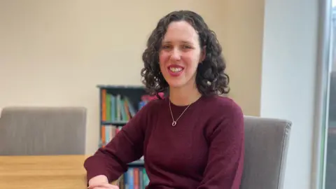 A woman with dark curly, shoulder-length hair looks into the camera. She is wearing a burgundy jumper with a silver necklace. She is sitting on a grey chair with a small bookcase in the background.
