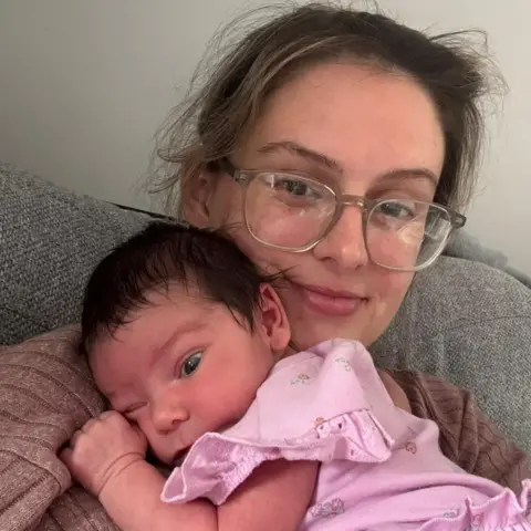 Chloe Chloe, a woman with light brown hair and clear rimmed glasses, sits with her daughter as a newborn baby lying on her and wearing a pink dress with frills.