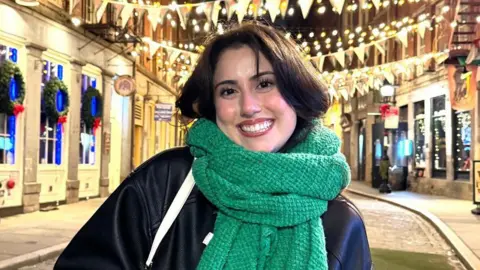 Samantha Rizzo Samantha smiles into the camera with a green scarf wrapped around her neck. She is wearing a black leather jacket and has brown hair. In the background we can see a festive street of shops lined with bunting and wreaths. It is early evening and there are yellow fairy lights switched on.