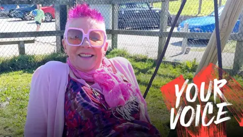 Annie Haslam Annie Haslam, with short pink spiky hair, pink glasses, pink scarf and pink cardigan, sits on a big swing outside
