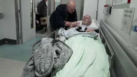 BBC A man leans over an elderly patient lying on a trolley in a hospital corridor, with other patients and equipment visible in the background.