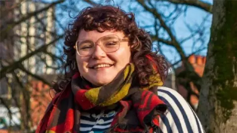 Tessa Marshall Tessa smiles at the camera. It is a head and shoulders shot of her, she has shoulder length brown curly hair, a nose stud, and wears glasses. She is wearing a yellow, red and black patterned scarf and a blue and white striped top. There are trees behind her, and a sky-scraper in the background. 
