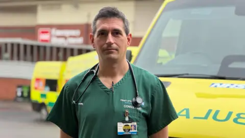 BBC A man with dark grey hair stands in green hospital scrubs by an ambulance, in front of a hospital building.