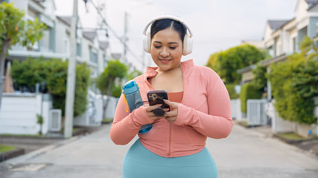 A women wearing headphones outdoors looking at a smartphone. 