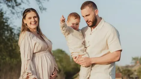 Dandelion & Rose Photography Chloe and her family all wear beige and stand outside in the sunshine with trees visible in the background as part of a professional family photoshoot. Chloe is visibly pregnant and holds her stomach smiling, while her partner holds their son.