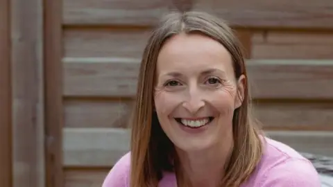 Caroline Stafford Close up of Caroline, in a pink jumper, smiling. There is a bare wooden fence behind her.