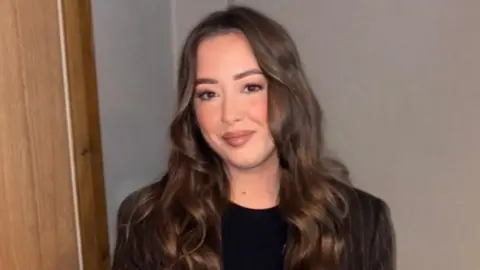 Laura Maguire Laura is a young woman with long, brown hair. She is smiling at the camera. She is standing against a white wall and is wearing a brown pinstripe blazer and a black t-shirt.