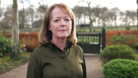 BBC Dr Susan Gilby wears a dark green shirt and is standing on a path in front of a gate with red flowers and tall trees in the background