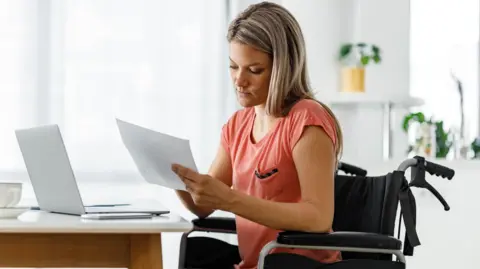 Getty Images A woman sitting in a wheelchair in front of her laptop, looking at paperwork.
