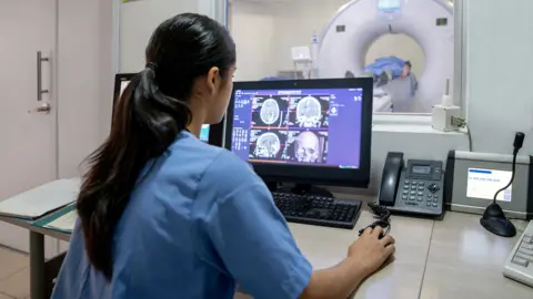 Getty Images Stock photo shows a person in a doctor's overall using a computer that shows brain scans, with a person being inserted into an MRI machine in the background in a hospital environment.