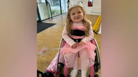 Charlie Brown Dani-Rae Brown is wearing a tiara and a pink princess costume, and holding a wand, while sitting in a wheelchair in a room indoors.