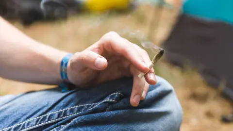 Getty Images A close-up stock photo that shows the knee and hand of a man wearing jeans sitting cross legged.  He's holding what appears to be a half-smoked cannabis joint.