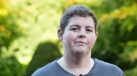 BBC Abbie Harvey is stood in the gardens of the Parkland Place Rehabilitation Centre with green trees and bushes behind her. She's wearing a grey t-shirt and is looking directly at the camera.