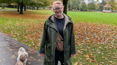 Cathy Read Woman in green jumper with leaves behind her and a small dog at her side. She is smiling at the camera