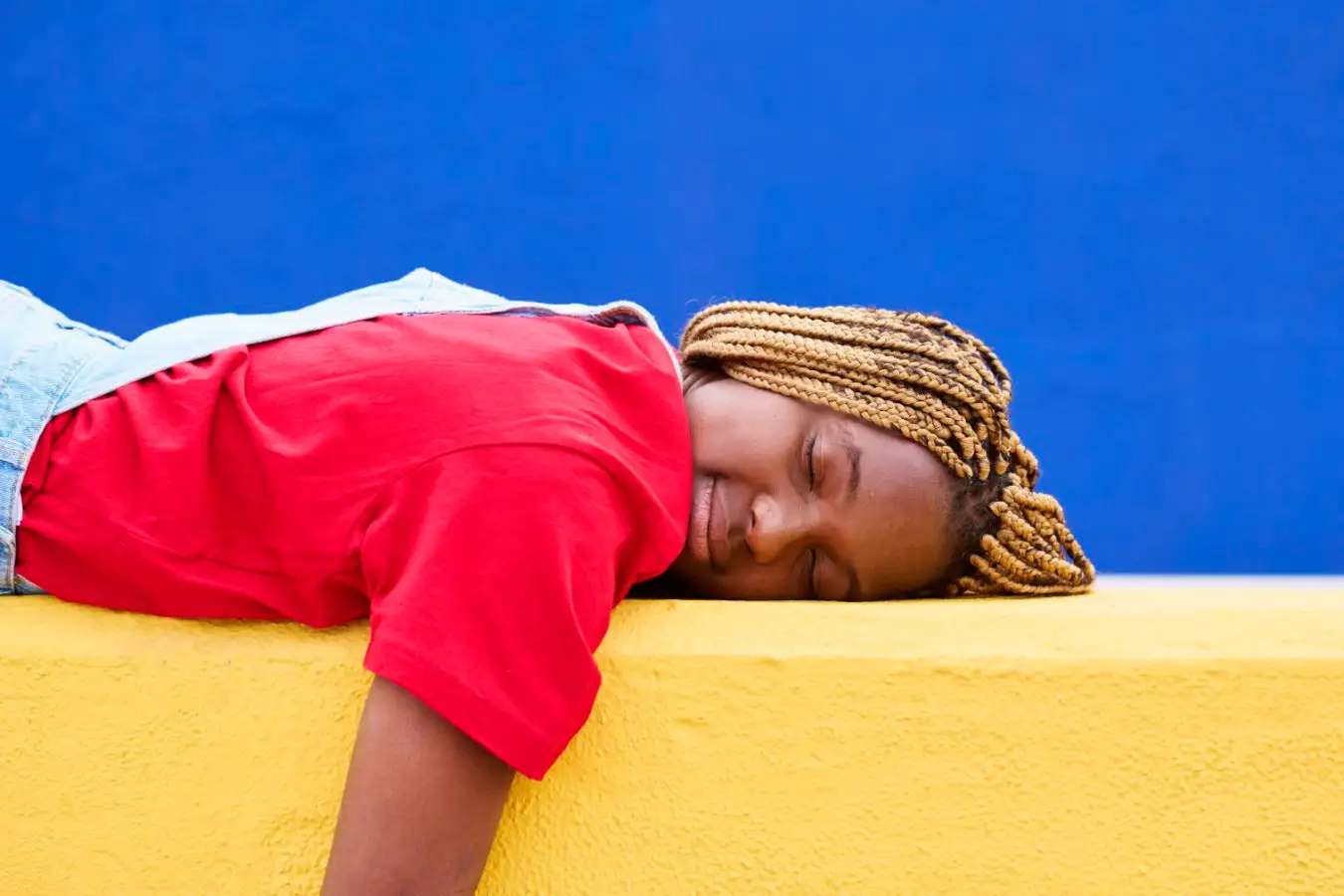 pretty colored woman in casual clothes, lying on a yellow colored wall with her eyes closed, resting