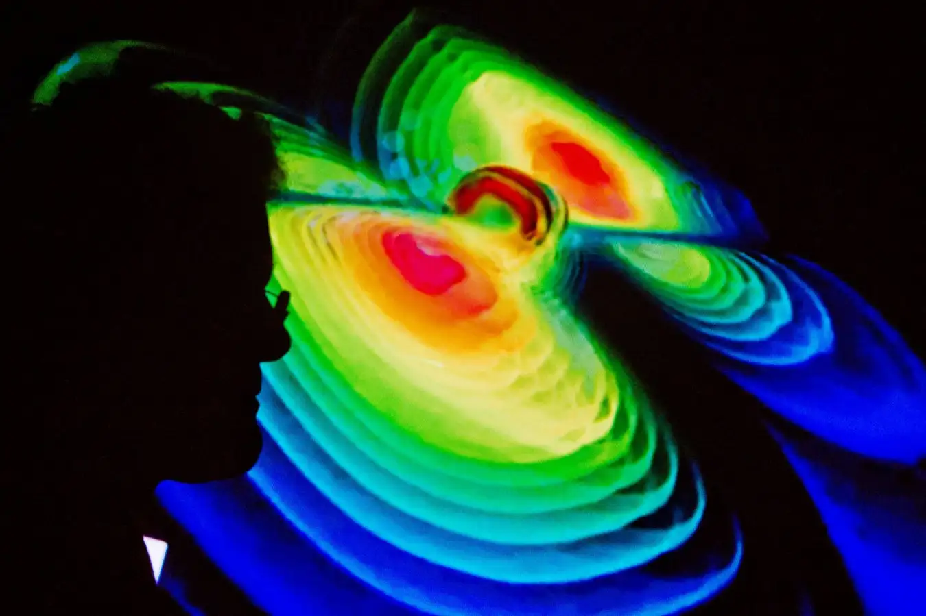 The silhouette of a scientist against a visualisation of gravitational waves - depicted as concentric rainbow waves 