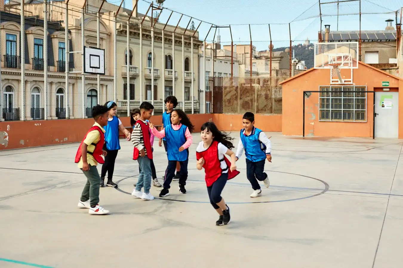 Elementary age boys and girls wearing red and blue scrimmage vests playing together on schoolyard sports court.