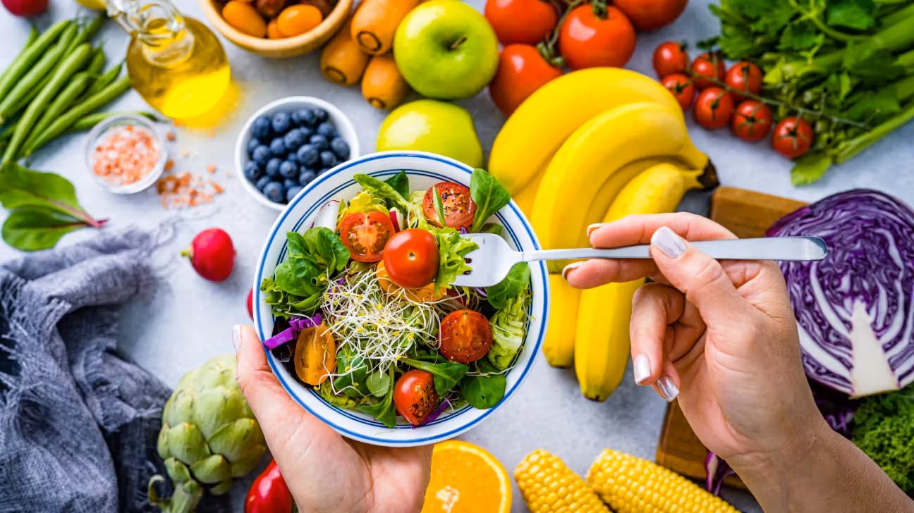 A person holding a fork in a bowl of salad surrounded by colorful fruits and vegetables